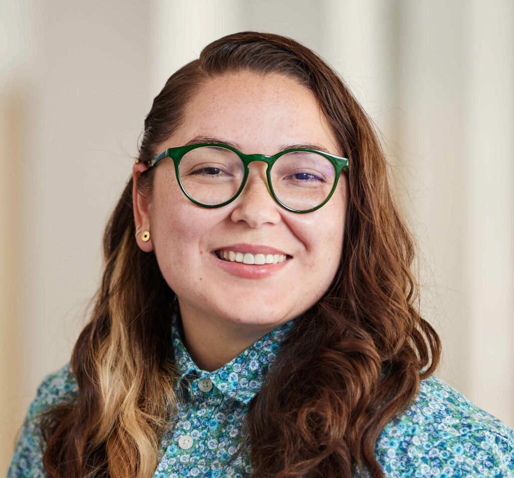 A headshot of a female with black round glasses smiling at the camera. She is wearing a blue flower patterned button up and her hair is wavy brown with a streak of blonde on the left side.