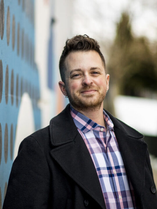 This is headshot of a man smiling at the camera. He is wearing a pink, blue, and white flannel with a black trench coat.