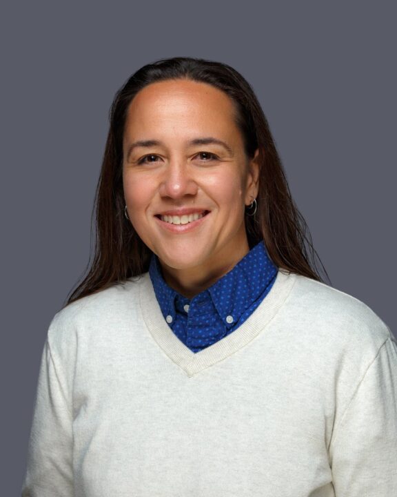 This is a headshot of a woman that has silver hoop earrings. She is also wearing a blue collar shirt with a white sweater over it.