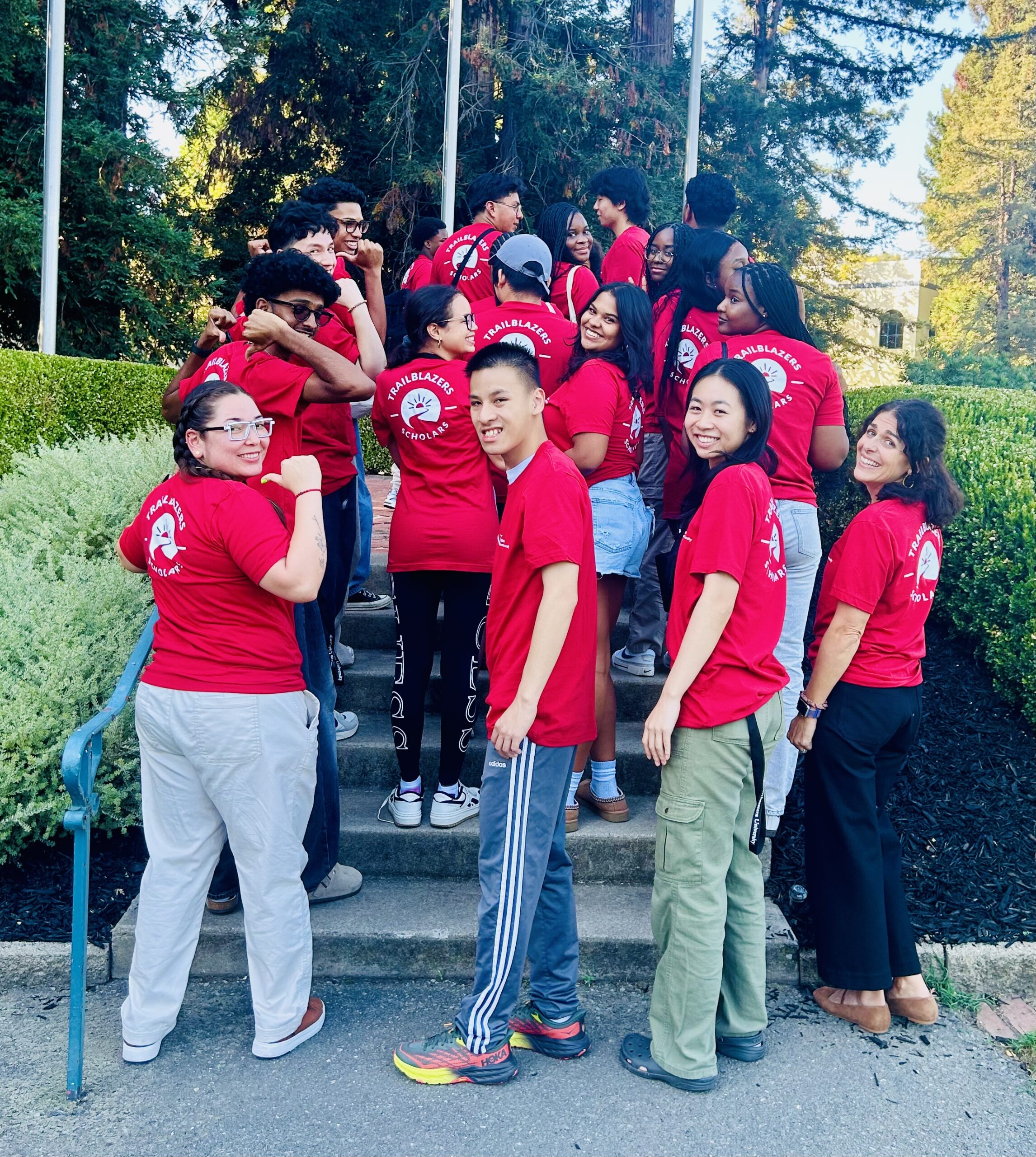 A group of students wearing red t-shirts from Northeastern University. They are gathered on the stairs and have their backs towards the cameras while looking at the camera smiling. There are large trees in the background and bushes on each side of the stairs.