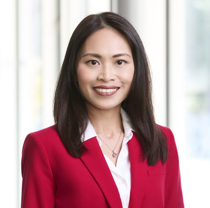 A headshot of an Asian woman with chestnut brown hair smiling at the camera. She is wearing a red suit and a white top.