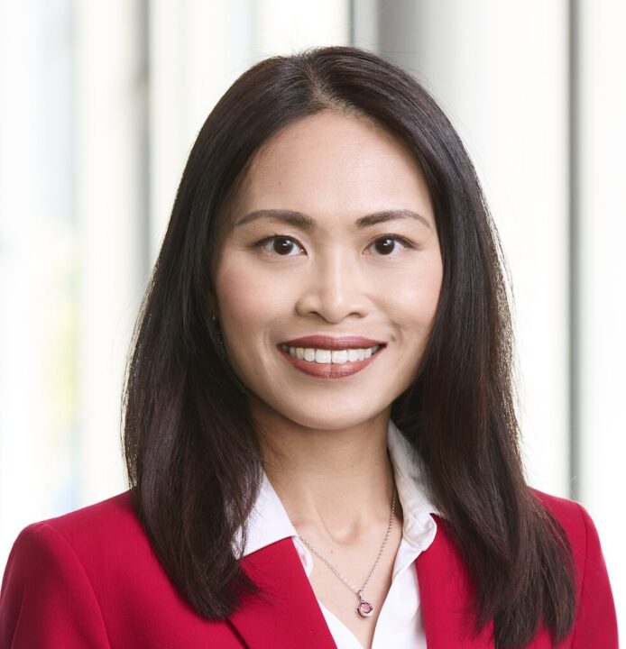 A headshot of an Asian woman with chestnut brown hair smiling at the camera. She is wearing a red suit and a white top.