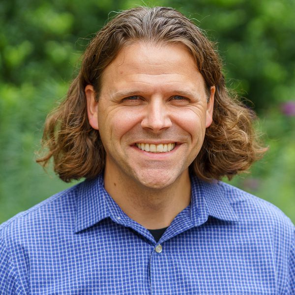 A caucasian man in a buttoned up blue shirt. He is smiling and has brown hair. 